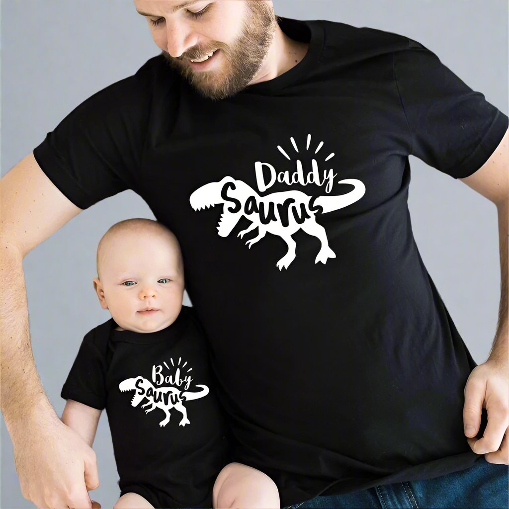 Father and baby wearing matching dinosaur-themed shirts, black shirts with 'Daddy Saurus' and 'Baby Saurus' designs