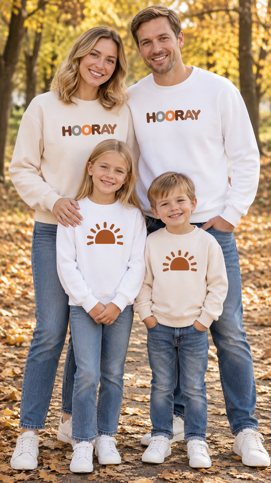 Cream adult crewneck sweatshirt printed HOORAY hanging beside a child's sweatshirt with a rust-orange half sun.